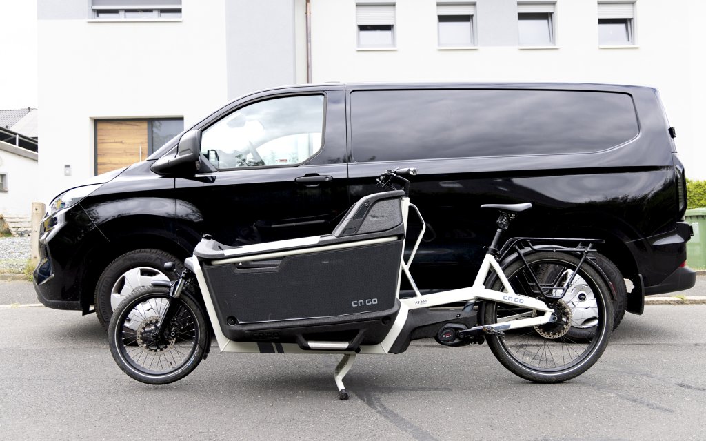 Weißes Cargobike steht vor einem schwarzen Lieferwagen.