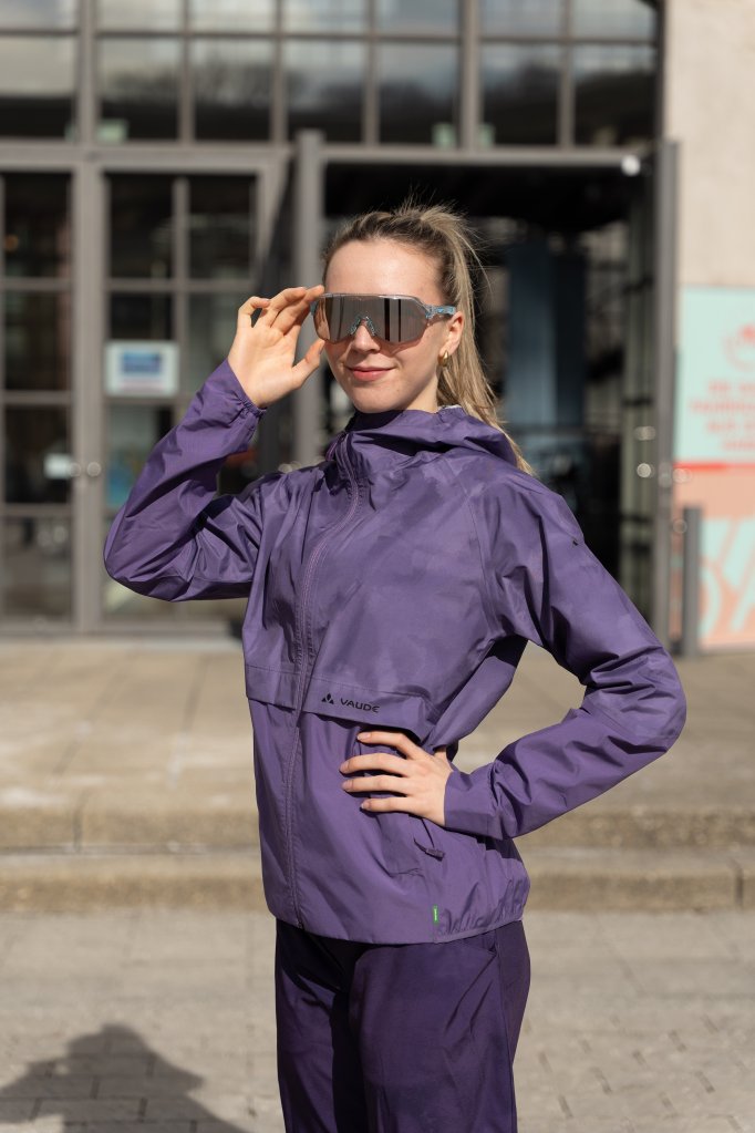 Frau steht mit Sonnenbrille und Fahrradjacke in der Sonne.