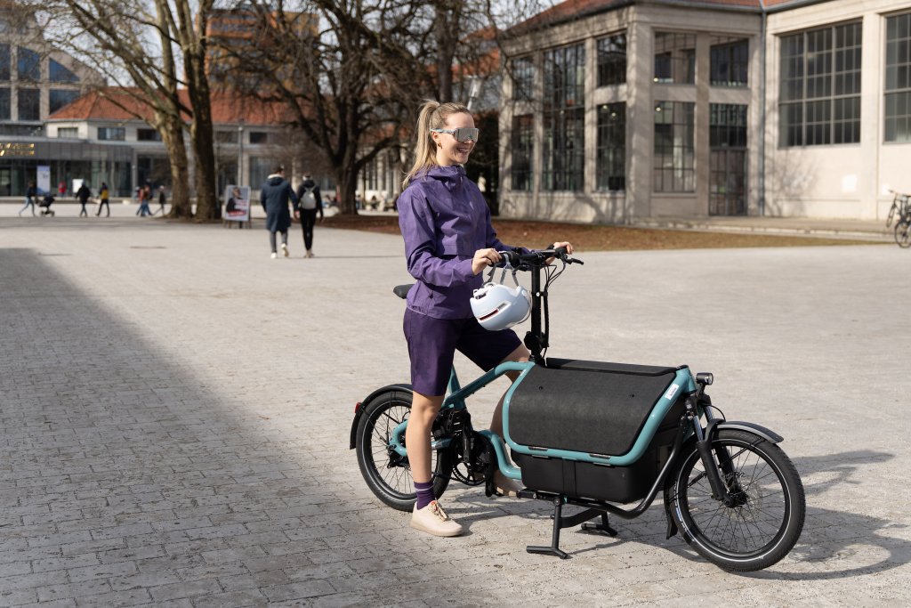 Frau steht mit Lastenrad auf Platz in der Sonne.