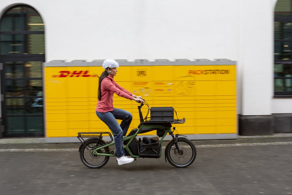 Frau fährt mit Lastenrad an Poststation vorbei.