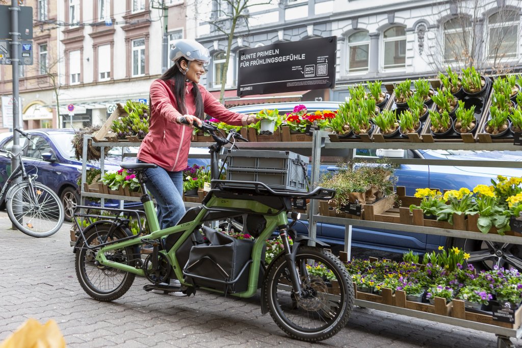 Frau steht mit Lastenrad vor Blumen.
