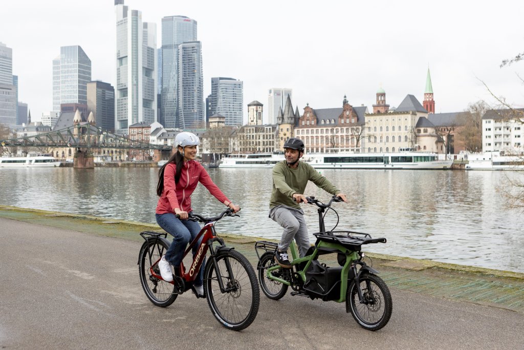 Frau auf E-Bike und Mann auf Lastenrad fahren den Main in Frankfurt entlang.
