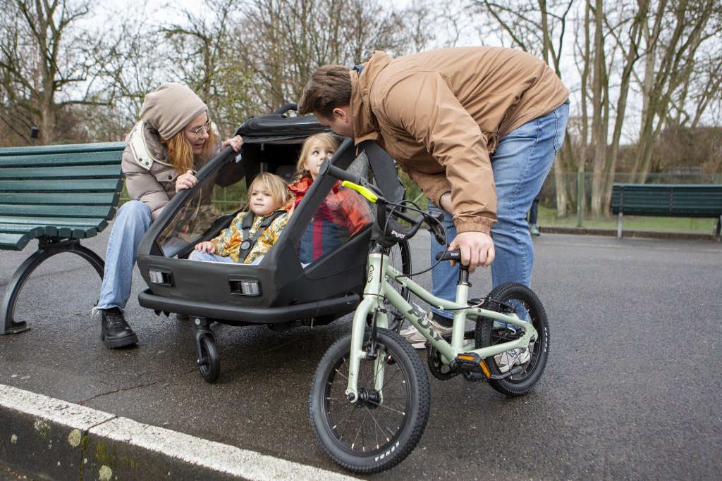 Familie mit zwei Kindern steht mit Kinderanhänger und Kinderrad an einer Parkbank.
