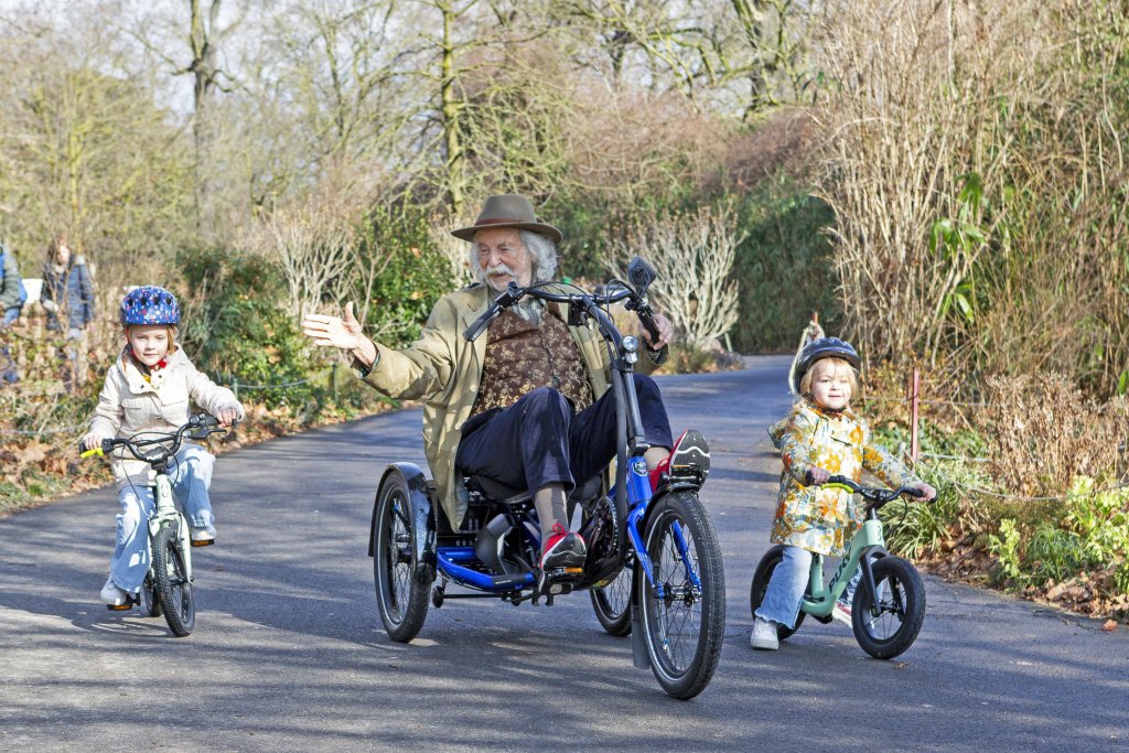 Älterer Mann mit Sesseldreirad fährt mit Kindern auf Laufrad und Fahrrad durch einen Park.