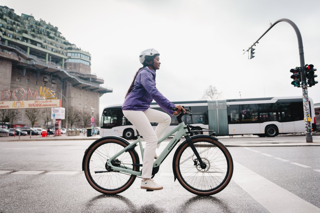 Frau fährt mit E-Bike durch die Stadt. Im Hintergrund fährt ein Bus.