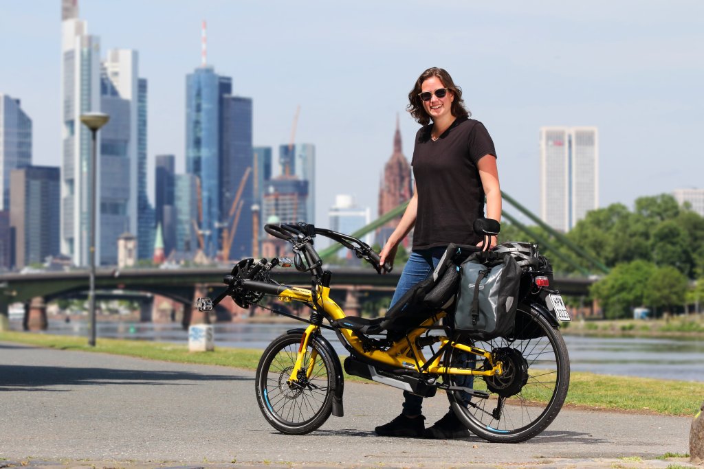Eine Frau steht mit einem gelben Liegerad mit Versicherungskennzeichen am Gepäckträger vor der Skyline einer Großstadt.