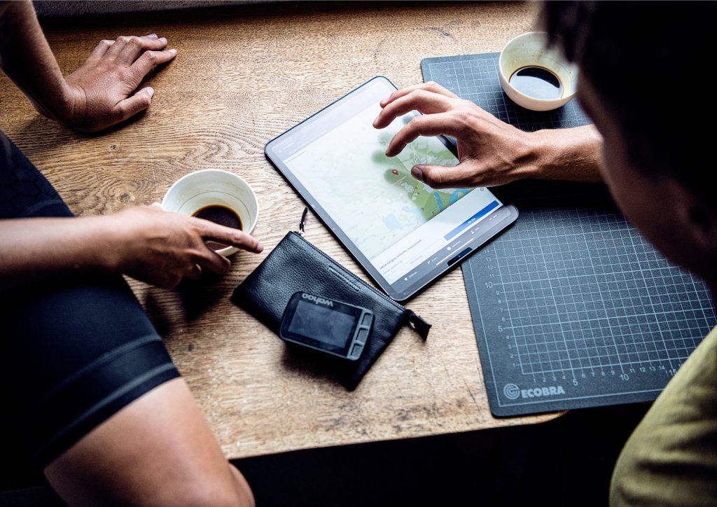Ein Tablet liegt zwischen zwei Kaffeetassen und einem Navigationsgerät für Fahrräder auf einem Tisch. Zwei Personen beschäftigen sich mit einer Kartenanwendung darauf.
