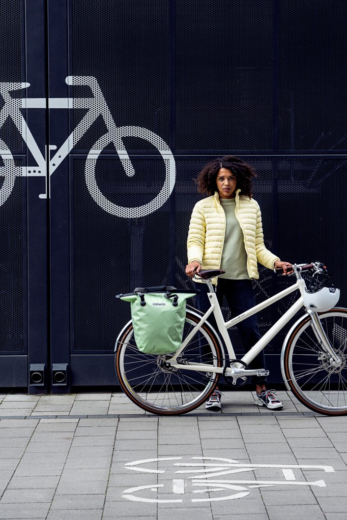 Eine Frau steht mit einem weißen Fahrrad vor einem dunkelblauen Metalltor mit einem Fahrradsymbol.