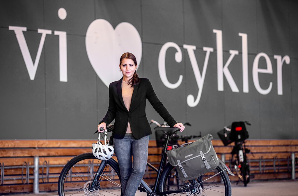 Eine Frau steht an einem Fahrrad gelehnt mit einem Helm am Lenker hängend. Im Hintergrund sind Fahrradständer zu erkennen und ein großes weißes Herz an einer grauen Wand mit dem Wort ?cykler? daneben.