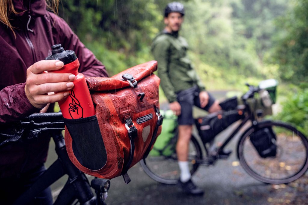 Eine Person nimmt eine Trinkflasche aus einem seitlichen Einschubfach an einer Fahrradlenkertasche.