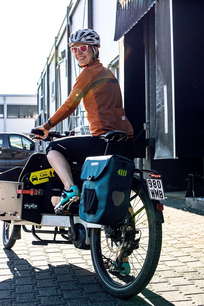 Eine Person auf einem Lastenrad schaut über die Schulter zur Kamera und lächelt.