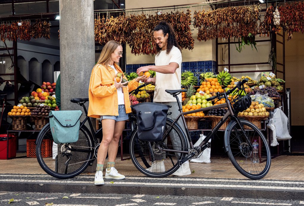 Eine Frau und ein Mann stehen mit Fahrrädern vor einem Obststand am Straßenrand und packen Obst in Fahrradtaschen.