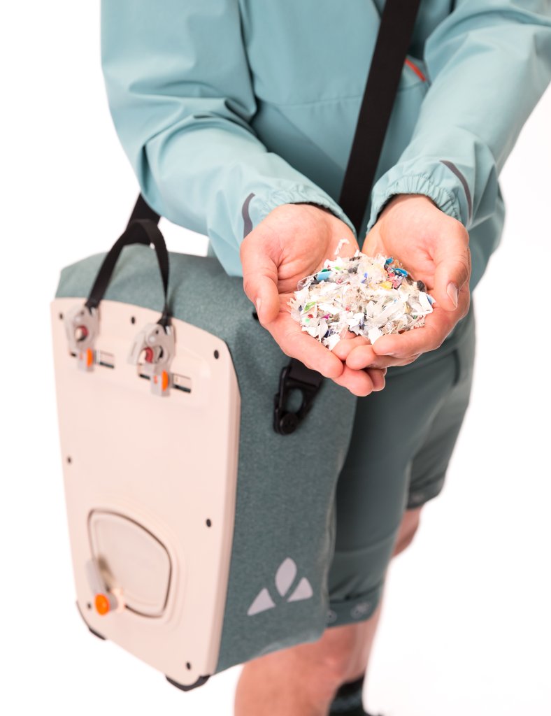 Eine Person hält Schnipsel von Kunststoffabfällen vor sich und trägt über der Schulter hängend eine Fahrradtasche.