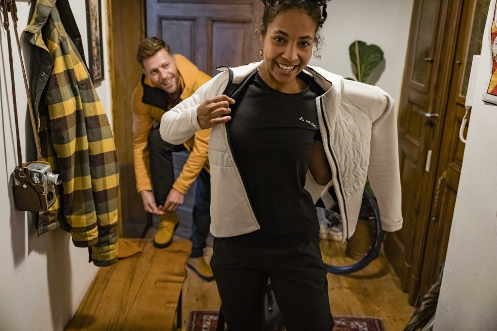 Eine Frau zieht sich im Flur einer Wohnung eine Jacke an. Ein Mann im Hintergrund zieht sich Schuhe an.
