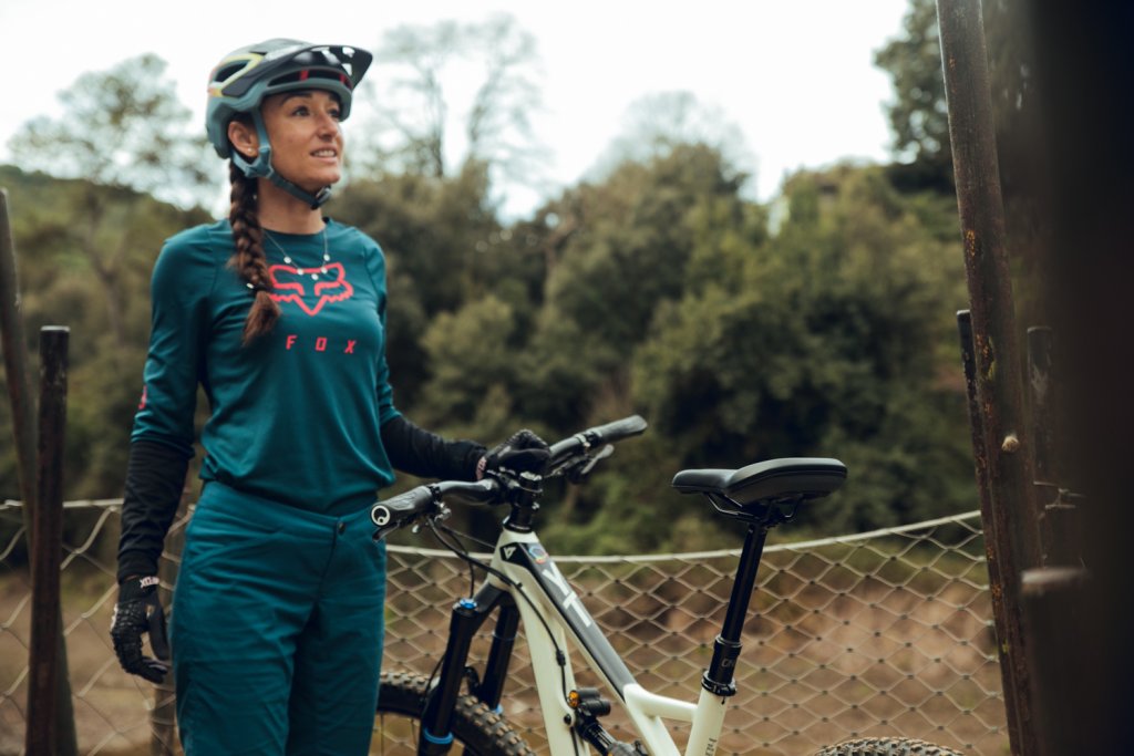 Eine Frau steht vor einem Zaun vor Bäumen und hält mit der linken Hand ein Mountainbike am Lenker.