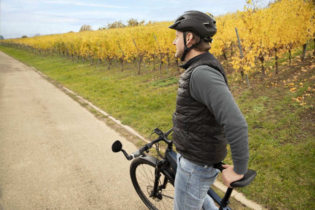 Ein Mann steht mit einem Fahrrad neben einem herbstlich gefärbten Weinhang auf einem Asphaltweg