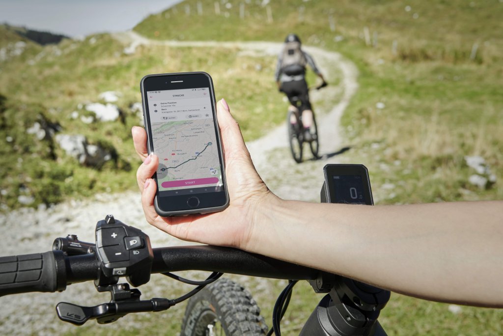 Eine Person hät ein Smartphone mit Karten-App darauf über den Lenker eines E-Bikes. Im Hintergrund fährt eine Person einen Wirtschaftsweg entlang.