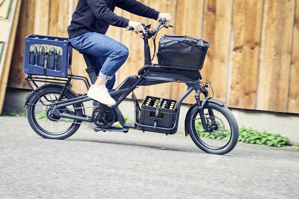 Mensch fährt beladenes Cargobike vor Holzwand.