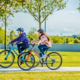 Zwei Kinder fahren auf Fahrrädern einen Betonweg zwischen Rasenflächen entlang.
