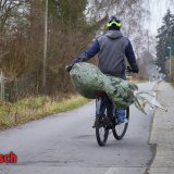 Eine Person fährt mit einem verpackten Weihnachtsbaum quer auf dem Gepäckträger eines Fahrrades und hält ihn dabei mit einer Hand am Stamm fest. Darunter steht in roten Bruchstaben 