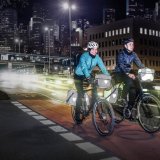 Zwei Personen auf Fahrrädern mit Licht und reflektierenden Elementen an den Fahrradtaschen vor vielen Lichtern einer Großstadt.