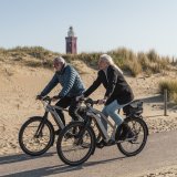 Ein Mann und eine Frau fahren auf E-Bikes einen Radweg vor einer Sanddüne entlang.
