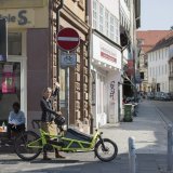Eine Frau steht neben einem Lastenrad am Eingang einer innerstädtischen Einbahnstraße und zeigt auf das Verkehrsschild dazu. Ihr Fahrradhelm hängt am Lenker.