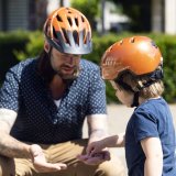Ein Kind nimmt etwas aus der Hand eines Mannes, der vor ihm hockt. Beide tragen orange Fahrradhelme.