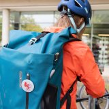Blick auf einen Rucksack, getragen von einer Radfahrerin, die scheinbar gerade ihr Fahrrad abstellt.