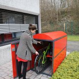 Eine Frau stellt ein Fahrrad in einem roten Metallkasten mit vorne geöffneter Tür ? einer sogenannten Fahrradgarage ? ab.