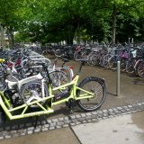 Ein Lastenrad mit einem Faltrad auf der Ladefläche, abgestellt am Rande einer großen Stellfläche für Fahrräder.