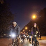Eine Gruppe Radfahrer:innen mit deutlich sichtbarer Beleuchtung fährt bei Dunkelheit auf einer Straße auf die Kamera zu.