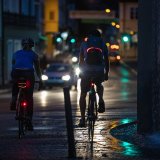 Zwei Personen fahren nachts auf Fahrrädern durch eine Innenstadt. An ihren Fahrrädern sind Rücklichter zu sehen und an ihren Rucksäcken und Taschen leuchtende oder reflektierende Elemente.