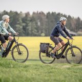 Eine Frau und ein Mann fahren auf E-Bikes zwischen Feldern und Wiesen entlang.