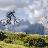 Eine Person springt mit einem E-Mountainbike. Im Hintergrund ist eine alpine Bergkette zu erkennen.