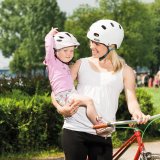 Eine Frau steht neben einem Fahrrad und lächelt das Kind an, das sie auf dem Arm trägt. Beide tragen einen Fahrradhelm.