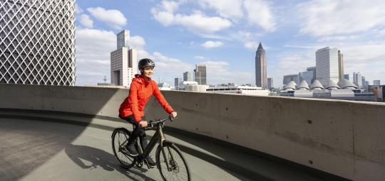 Eine Frau fährt mit einem E-Bike vor der Skyline von Frankfurt entlang.