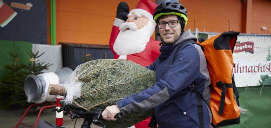 Ein Mann steht mit einem E-Bike vor einer Verkaufsstelle für Weihnachtsbäume. Einen verpackten Weihnachtsbaum hat er auf Lenker und Sattel abgelegt.