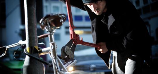 Ein Mann setzt einen Bolzenschneider an einem Bügelschloss an, mit dem ein Fahrrad an einem Metallpfosten gesichert ist.