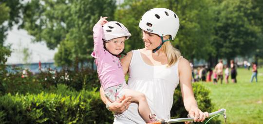 Eine Frau steht neben einem Fahrrad und lächelt das Kind an, das sie auf dem Arm trägt. Beide tragen einen Fahrradhelm.