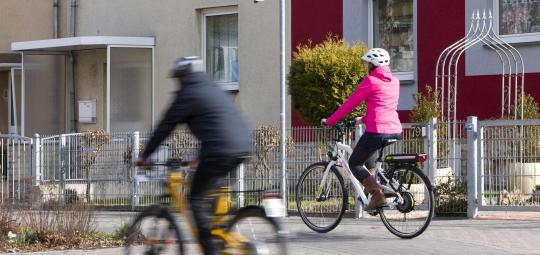 Zwei Personen fahren in dieselbe Richtung auf Fahrrädern durch die Stadt ? eine auf der Fahrbahn und eine auf dem geteilten Fuß- und Radweg.