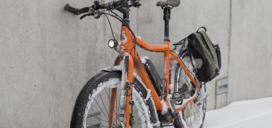 Ein orangefarbenes E-Bike mit Schnee in den Rädern und am Rahmen steht an eine Wand gelehnt.