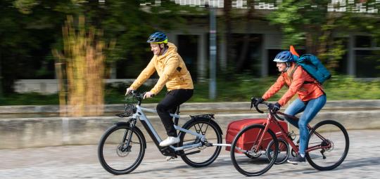 Ein Mann auf einem E-Bike mit Anhänger und eine Frau auf einem Rennrad fahren nebeneinander einen gepflasterten Weg entlang.