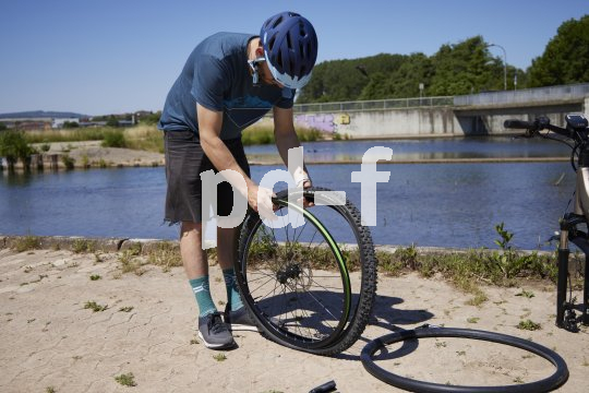Ein Radfahrer steht vor einem Gewässer und zieht einen Fahrradreifen auf ein Vorderrad. Am Boden daneben liegt ein leicht aufgepumpter Fahrradschlauch.