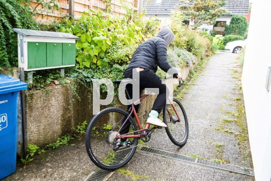 Eine Person mit Kapuze steigt zwischen Briefkästen und einer Hauswand auf ein Fahrrad, um loszufahren.