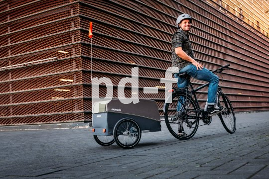 Ein Mann steht mit einem Fahrrad mit Anhänger auf einer gepflasterten Fläche und schaut lächelnd nach hinten.