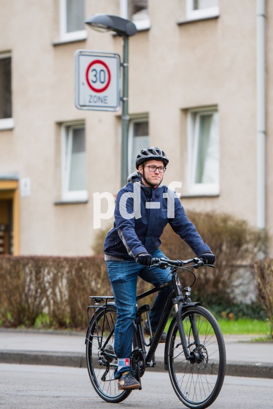 Ein Mann fährt auf einem Fahrrad auf einer Straße. Im Hintergrund ist vor einem Wohnhaus ein Verkehrsschild mit Geschwindigkeitsbegrenzung 30 km/h zu sehen.