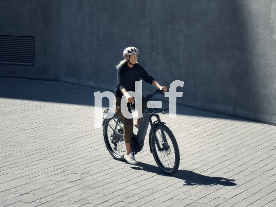 Frau fährt mit E-Bike auf Pflasterweg vor Betonwand.