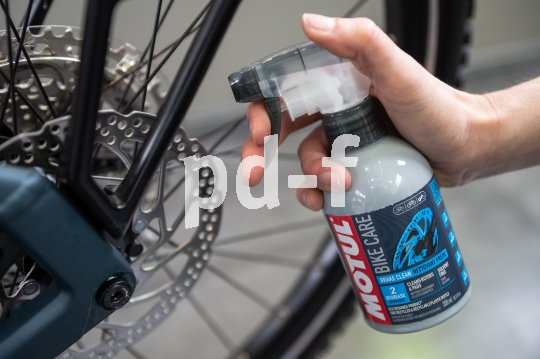 Eine Person richtet eine Sprühflasche mit Bremsenreiniger auf die Bremsscheibe eines Fahrrades.