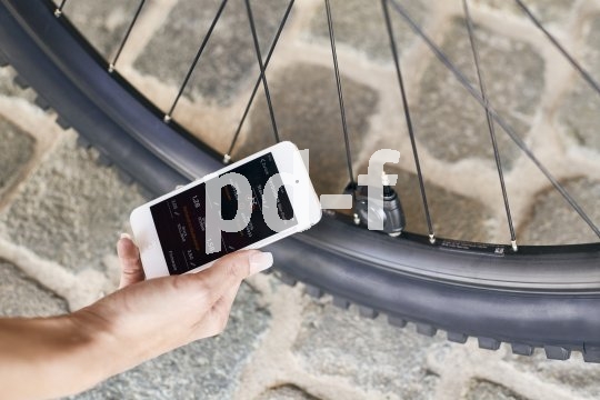 Eine Person hält ein Smartphone vor einen Fahrradreifen, an dessen Ventil ein kleines, schwarzes Gerät montiert ist.